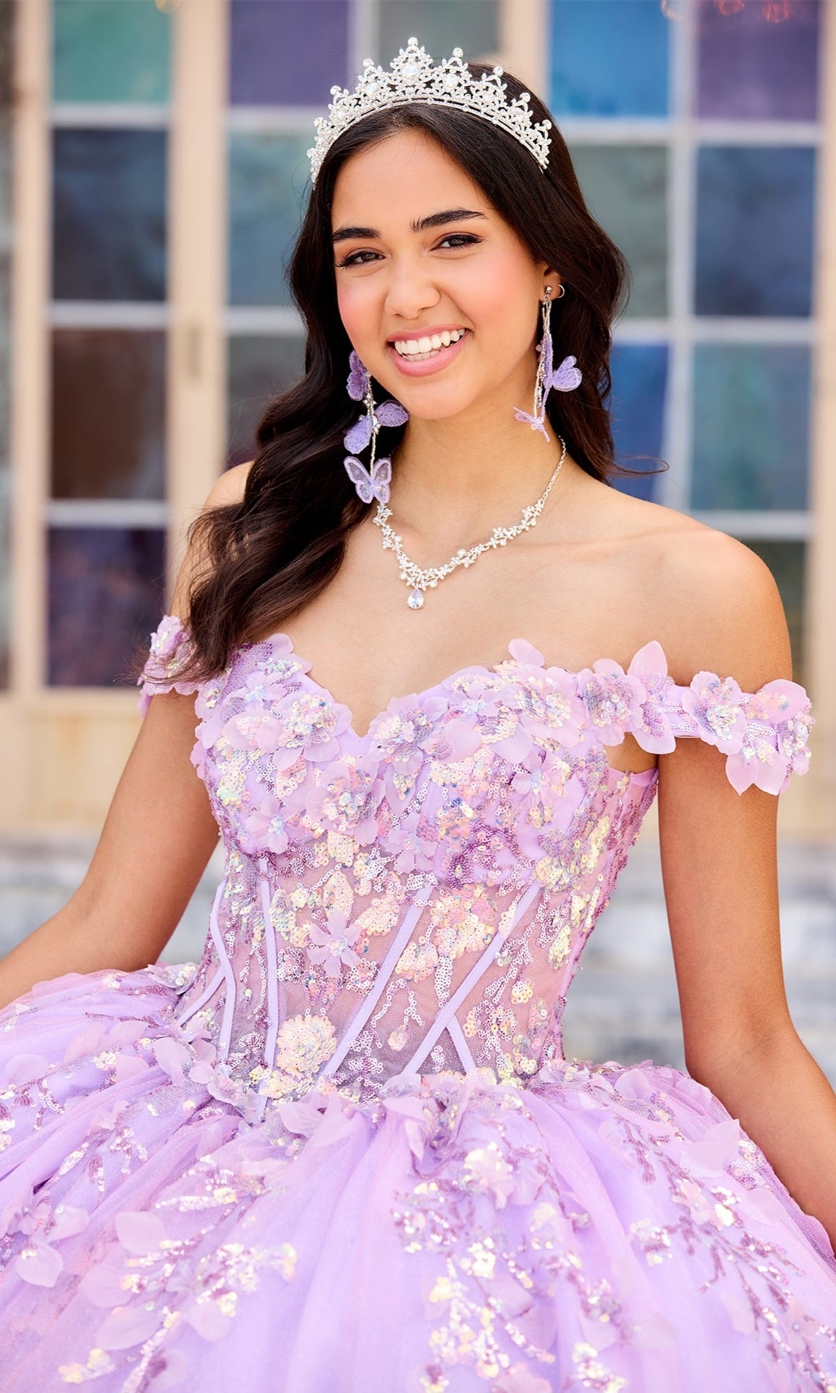 A young woman wearing the Quinceanera Dress PR30162 By Princesa, featuring a lavender floral ombre design and off-the-shoulder neckline, smiles in front of a windowed background.